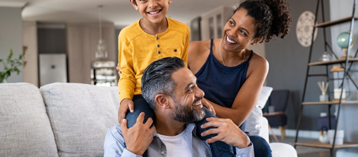 Cheerful african mother and indian father playing with son at home. Cute boy enjoying sitting on father shoulder while looking at camera. Middle eastern family having fun together on the sofa at home.